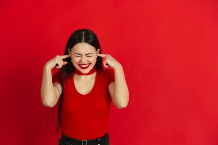 Crazy covering ears with hands. Caucasian young womans monochrome portrait isolated on red studio background. Beautiful female brunette model. Concept of human emotions, facial expression, sales, ad.の写真素材