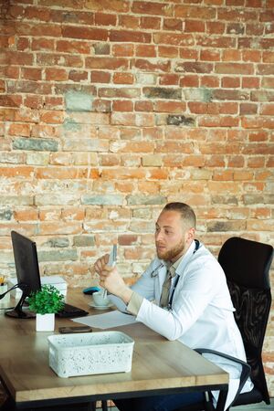 Doctor consulting for patient, working with laptop. Caucasian doctor during his work with patients, explaining recipes for drug, consulting. Daily hard work for health and lives saving during epidemic.の写真素材