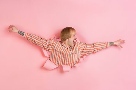 Crazy greeting. Cheerful caucasian young man poses in torn coral paper background, emotional expressive. Breaking on, breakthrought. Concept of human emotions, facial expression, sales, ad.の写真素材