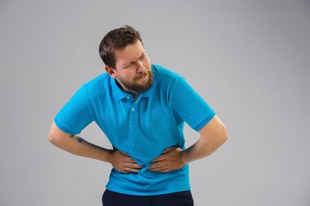 Young caucasian man suffers from abdominal pain, feels sick, ill and weakness isolted on gray studio background. Symptoms of desease or unhealthy lifestyle. Sickness, ilness, sad and upset. Copyspace.の写真素材