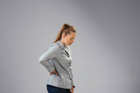Young caucasian woman suffers from back pain, feels sick, ill and weakness isolted on gray studio background. Symptoms of desease or unhealthy lifestyle. Sickness, ilness, sad and upset. Copyspace.の写真素材