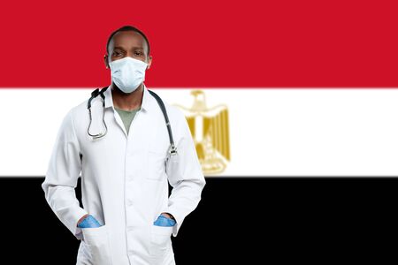 Young doctor with stethoscope and face mask praying for God with Egypt national flag on background. Looks sad, serious. Concept of healthcare and medicine, coronavirus pandemic, danger epidemic, COVID.の写真素材