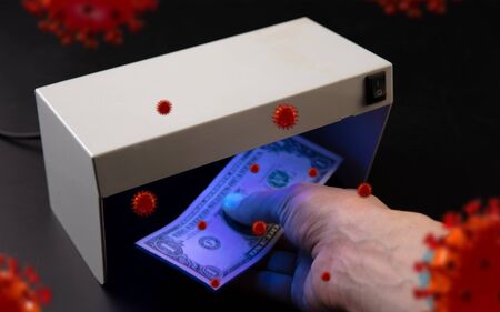 Male hands checking of banknotes in the detector, 3D models of coronavirus spreding around. The...