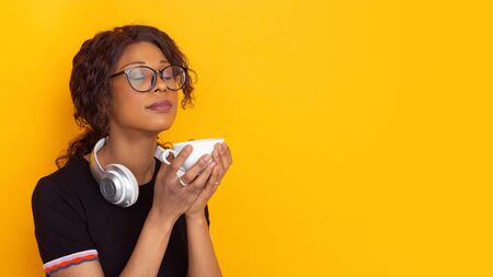 Joyful drinks coffee with eyes closed. African-american young womans portrait isolated on yellow background. Beautiful curly model. Concept of human emotions, facial expression, sales, ad, youth.の写真素材
