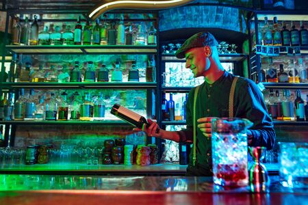 Professional barman finishes preparation of alcoholic cocktail for guest in multicolored neon light. Entertainment, drinks, service concept. Modern bar, crafted beverages, trendy neoned colors.の写真素材