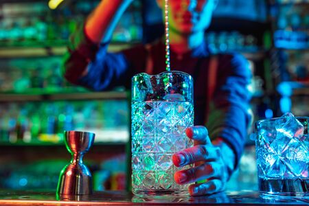 Close up of professional barman finishes preparation of alcoholic cocktail in multicolored neon light, gives it to client. Entertainment, drinks, service concept. Modern bar, trendy neoned colors.の写真素材
