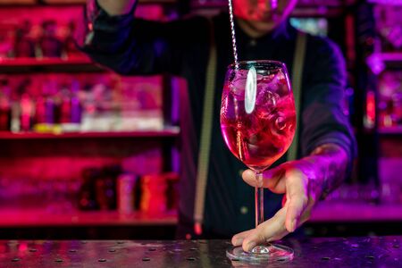 Close up of professional barman finishes preparation of alcoholic cocktail in multicolored neon light, gives it to client. Entertainment, drinks, service concept. Modern bar, trendy neoned colors.の写真素材