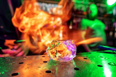 Close up big melting piece of ice on the bar counter in fireflames on it, preparation for a cocktail. Entertainment, drinks, service concept. Modern bar, trendy neoned colors. Bright, vibrant colors.の写真素材