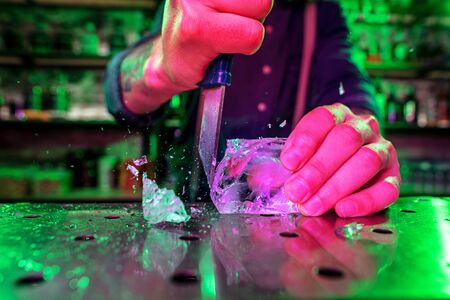 Close up of barman crushing a big piece of ice on the bar counter with a special bar equipment on it for a cocktail. Entertainment, drinks, service concept. Modern bar, trendy neoned colors.の写真素材
