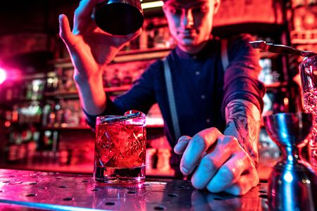 Close up of professional barman finishes preparation of alcoholic cocktail in multicolored neon light, gives it to client. Entertainment, drinks, service concept. Modern bar, trendy neoned colors.の写真素材