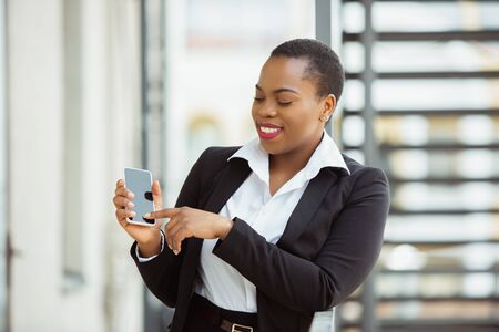 Using smartphone. African-american businesswoman in office attire smiling, looks confident and happy, busy. Finance, business, equality and human rights concept. Beautiful young feme model, successful.の写真素材