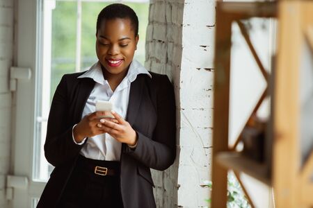 Using smartphone. African-american businesswoman in office attire smiling, looks confident and happy, busy. Finance, business, equality and human rights concept. Beautiful young feme model, successful.の写真素材