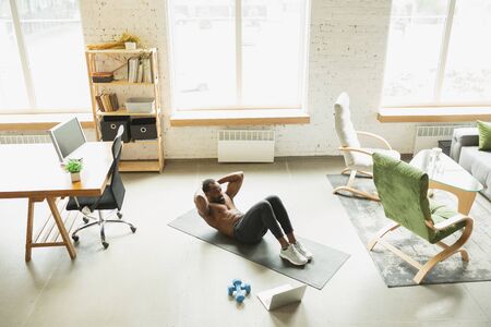 Young african-american man training at home during quarantine of coronavirus outbreak, doinc exercises of fitness, aerobic. Staying sportive during insulation. Wellness, movement concept. ABS workout.の写真素材