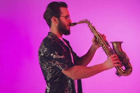 Young caucasian jazz musician playing the saxophone on gradient pink studio background in neon light. Concept of music, hobby, festival. Joyful, cheerful attractive guy. Colorful portrait of artist.の写真素材