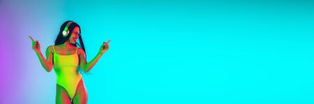 Beautiful young girl dancing with wireless headphones on gradient studio background in neon light. Woman in fashionable bodysuit. Facial expression, summer, weekend, beauty, resort concept. Vacations.の写真素材