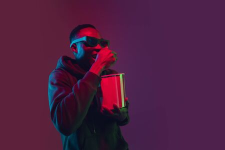Smiling. African-american mans portrait on gradient studio background in neon light. Beautiful male model with popcorn and 3D-eyewear. Concept of human emotions, facial expression, sales, ad, cinema.の写真素材