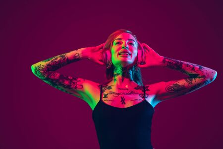 Listening to music, dancing with wireless headphones. Portrait of young caucasian woman on purple background in neon light. Human emotions, facial expression, sales, ad concept. Freaks culture.の写真素材