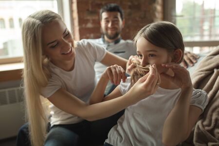 Mother, father and daughter at home having fun, comfort and cozy concept. Looks happy, cheerful and joyful. Beautiful caucasian family. Spending time together, doing a hairstyle, scrolling tablet.の写真素材