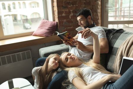 Mother, father and daughter at home having fun, comfort and cozy concept. Looks happy, cheerful, joyful. Beautiful caucasian family. Spending time together, reading books, using devices, taking selfie.の写真素材