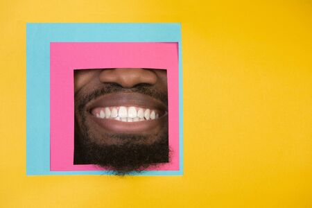 Smile. Mouth of emotional african-american man peeks throught square in yellow background. Trendy geometrical style, copyspace. Vibrant colors. Sales, proposal, finance and business concept. Framing.の写真素材