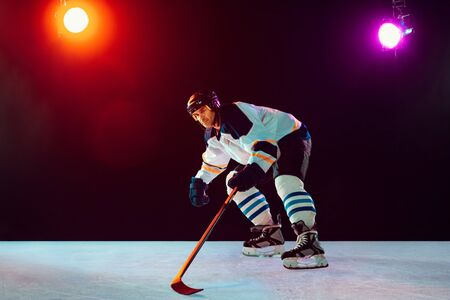 Champion. Male hockey player on ice court and dark neon colored background with flashlights. Sportsman in equipment, helmet practicing. Concept of sport, healthy lifestyle, motion, wellness, action.の写真素材