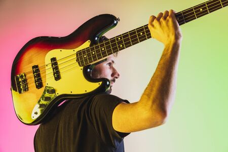 Close up young and joyful caucasian musician playing bass guitar on gradient studio background in neon light. Concept of music, hobby, festival. Colorful portrait of modern artist. Inspired improvising.の写真素材