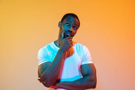 Thoughtful, thinking. African-american mans modern portrait on gradient orange studio background in neon light. Beautiful afro model. Concept of human emotions, facial expression, sales, ad.の写真素材