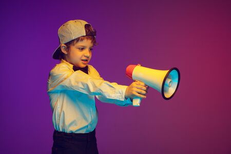 Shouting with megaphone, emotional. Joyful caucasian boys portrait on dark studio background in neon...