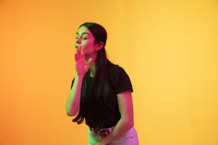 Whispering a secret. Caucasian young womans portrait on yellow studio background in neon. Beautiful...