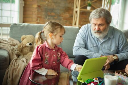 Grandfather and grandchild playing together at home. Happiness, family, relathionship, learning and education concept. Sincere emotions and childhood. Reading books, playing gadgets, look happy.の写真素材