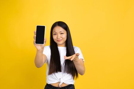 Showing blank phone screen. Portrait of young asian woman on yellow studio background. Stylish,...