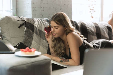 Drinking wine, reading magazine. Portrait of pretty young girl in modern apartment in the morning. Resting, calm, salisfied. Youth and wellness concept. Elegance woman in silk dress, comfortable.の写真素材