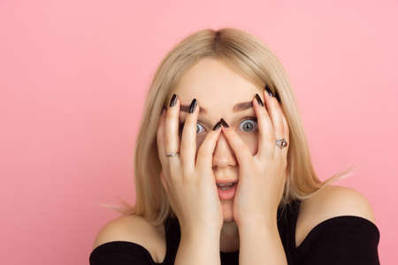Shocked hiding face. Portrait of young caucasian woman with bright emotions on coral pink studio background. Blonde female model. Concept of human emotions, facial expression, sales, advertising, youth.の写真素材
