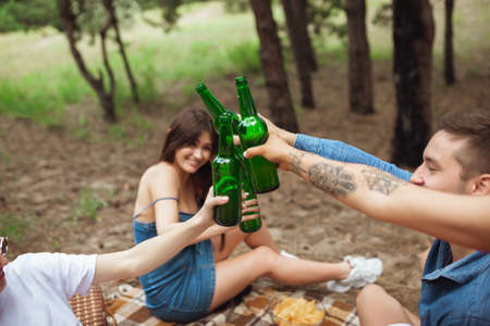 On the way. Group of friends clinking beer bottles during picnic in summer forest. Lifestyle, friendship, having fun, weekend and resting concept. Looks cheerful, happy, celebrating, festive.の写真素材