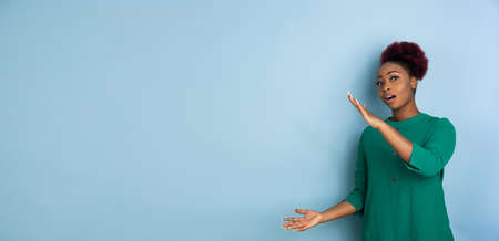 Holding blank copyspace. African-american young womans portrait isolated on blue studio background. Beautiful female model. Concept of human emotions, facial expression, sales, ad. Copyspace. Flyerの写真素材