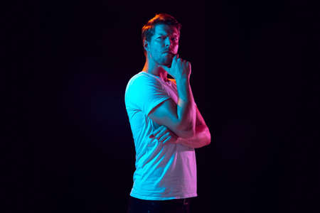 Thoughtful, serious. Caucasian young mans portrait on black studio background in neon light. Beautiful male model in white shirt. Concept of human emotions, facial expression, sales, ad. Copyspace.の写真素材