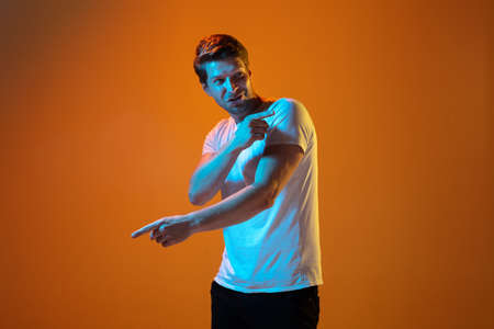 Pointing at side. Caucasian young mans portrait on gradient orange studio background in neon light. Beautiful male model. Concept of human emotions, facial expression, sales, ad. Copyspace.の写真素材