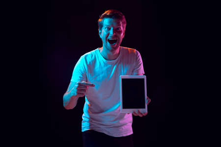 Pointing on tablet, crazy excited. Caucasian young mans portrait on black studio background in neon light. Beautiful male model. Concept of human emotions, facial expression, sales, ad. Copyspace.の写真素材