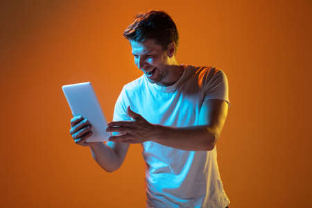 Laughting scrolling tablet. Caucasian young mans portrait on gradient orange studio background in neon light. Beautiful male model. Concept of human emotions, facial expression, sales, ad. Copyspace.の写真素材