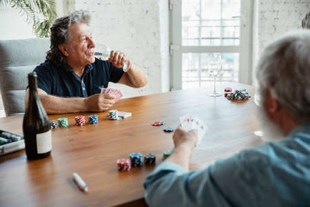 Young at heart. Two happy mature friends playing cards and drinking wine. Look delighted, excited. Caucasian men gambling at home. Sincere emotions, wellbeing, facial expression concept. Good old age.の写真素材