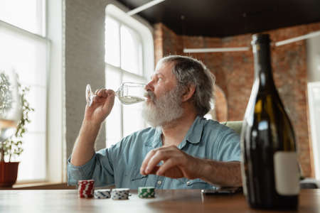 Exciting. Happy mature man drinking wine with friends during card game. Looks delighted, excited....