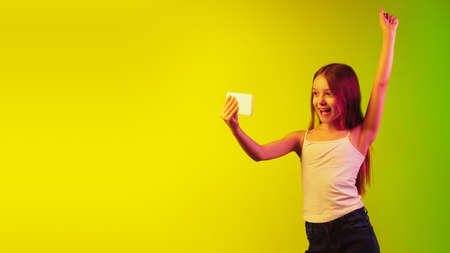 Playing phone excited. Little caucasian girls portrait on gradient studio background in neon light....