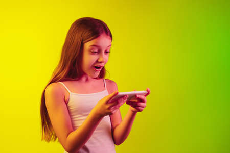 Playing phone excited. Little caucasian girls portrait on gradient studio background in neon light. Concept of human emotions, facial expression, modern gadgets and technologies, sales, ad. Copyspace.の写真素材
