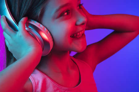 Listen to music with headphones, close up. Little girls portrait on gradient pink-blue background in...