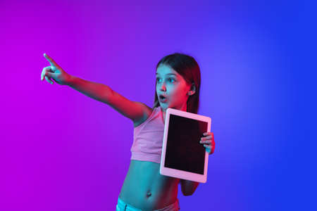 Pointing, showing tablet with blank screen. Little girls portrait on gradient pink-blue background in neon. Concept of human emotions, facial expression, modern gadgets and tech, sales, ad. Copyspace.の写真素材
