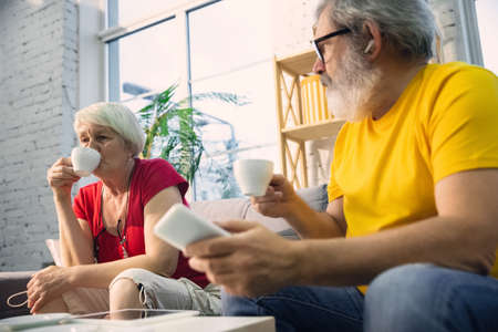 Couple of seniors smiling spending time together being quarantined - caucasians mature and retired man and woman using modern gadgets, talking, drinking tea. Eyes tired of gadgets life. Lifestyle.の写真素材