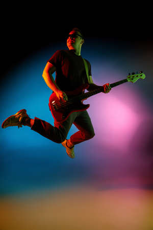 Jumping high. Young inspired and expressive musician, guitarist performing on gradient colored background in neon. Concept of music, hobby, festival, art. Joyful artist, colorful, bright portrait.の写真素材