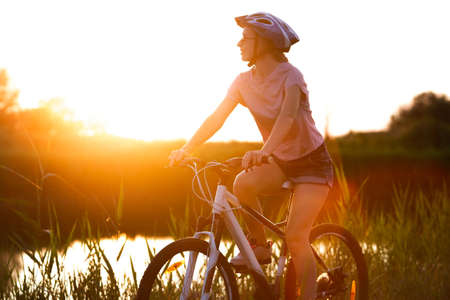 Nature. Joyful young woman riding a bicycle at the riverside and meadow promenade. Inspired by surrounded nature, summertime mood. Warm sunshine colors. Sport, activity, wellness, enjoyment concept.の写真素材