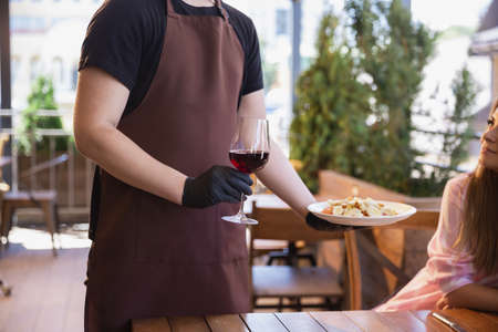 The waiter works in a restaurant in a medical mask, gloves during coronavirus pandemic. Representing new normal of service and safety. Putting the order, meals and drink, wine. Taking care of clients.の写真素材