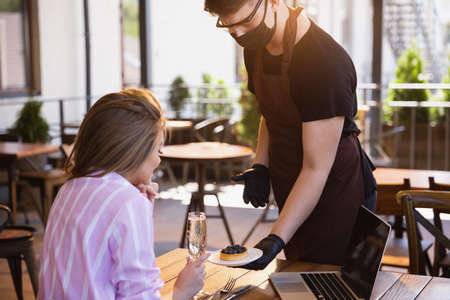 The waiter works in a restaurant in a medical mask, gloves during coronavirus pandemic. Representing new normal of service and safety. Giving dessert, tasty cake with berries. Taking care of clients.の写真素材